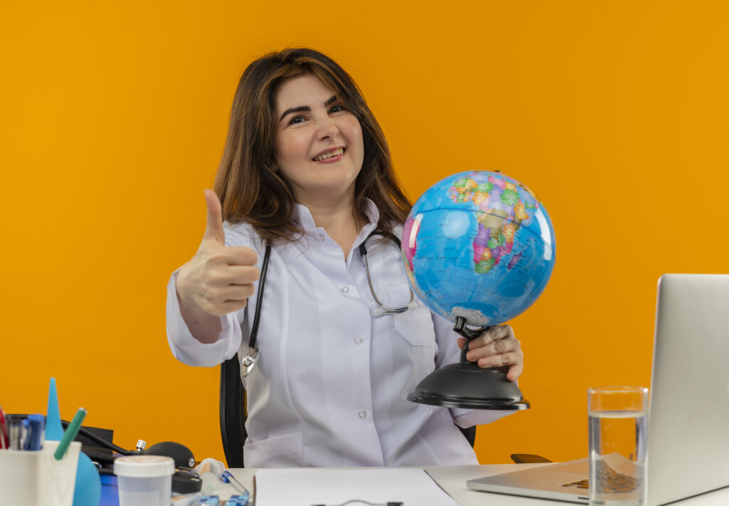 B2B Travel Agency: smiling middle-aged female doctor wearing medical robe with stethoscope sitting at desk work on laptop with medical tools holding globe her thumb up on isolated orange background with copy space