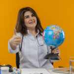 B2B Travel Agency: smiling middle-aged female doctor wearing medical robe with stethoscope sitting at desk work on laptop with medical tools holding globe her thumb up on isolated orange background with copy space