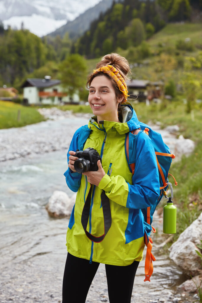 Pakt Travel Backpack:
Travel and outdoor activities concept. Optimistic lovely female hiker walks by small mountain stream, holds professional photocamera, makes photos of landscape, green forest and small house behind