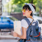 worst countries to visit: Lightly tanned brunette girl wears trendy accessories perplexedly looking around, holding city map. Stunning black-haired traveler carrying big blue backpack walking down the street with cars.