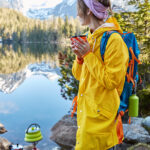 Things to do in Blue Ridge GA: Outdoor vertical shot of pensive female hacker holds a hot beverage in a teacup, making a drink on special tourist equipment, wearing a yellow raincoat, and rubber boots, admiring the scenic lake scape and mountains