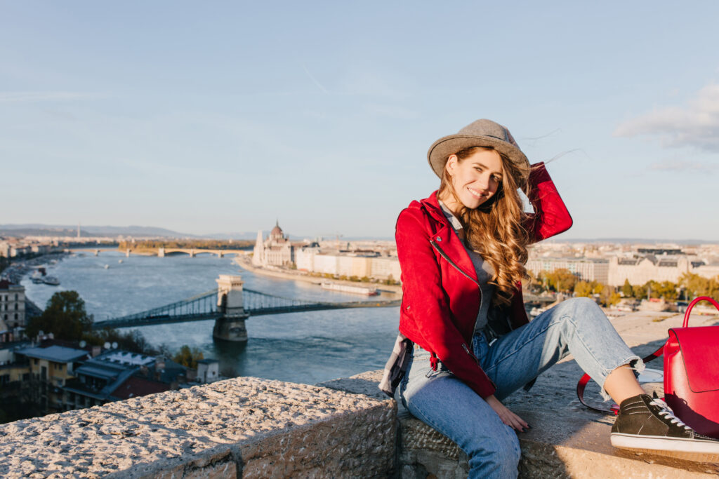 LAX to Europe:
Romantic girl in black gumshoes sitting on roof and playing with brown hair. Outdoor portrait of dreamy woman in red jacket posing with river on background..