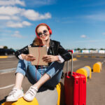 flights from Philadelphia to Atlanta: Smiling girl in sunglasses and headphones happily listening music on tablet with red suitcase near on road of airport