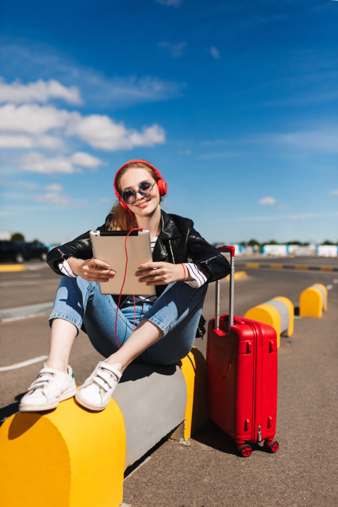 flights from Philadelphia to Atlanta: Smiling girl in sunglasses and headphones happily listening music on tablet with red suitcase near on road of airport