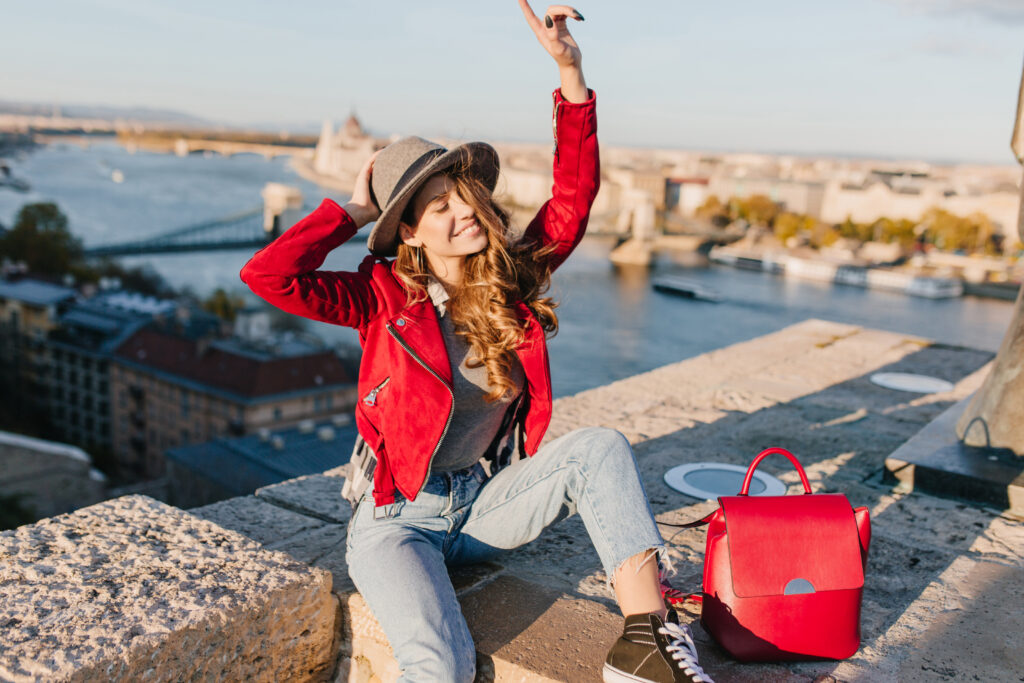Solo Travel Italy: Girl in elegant hat and vintage pants funny posing on river background. Outdoor portrait of graceful white woman in red jacket having fun during european trip in weekend.
