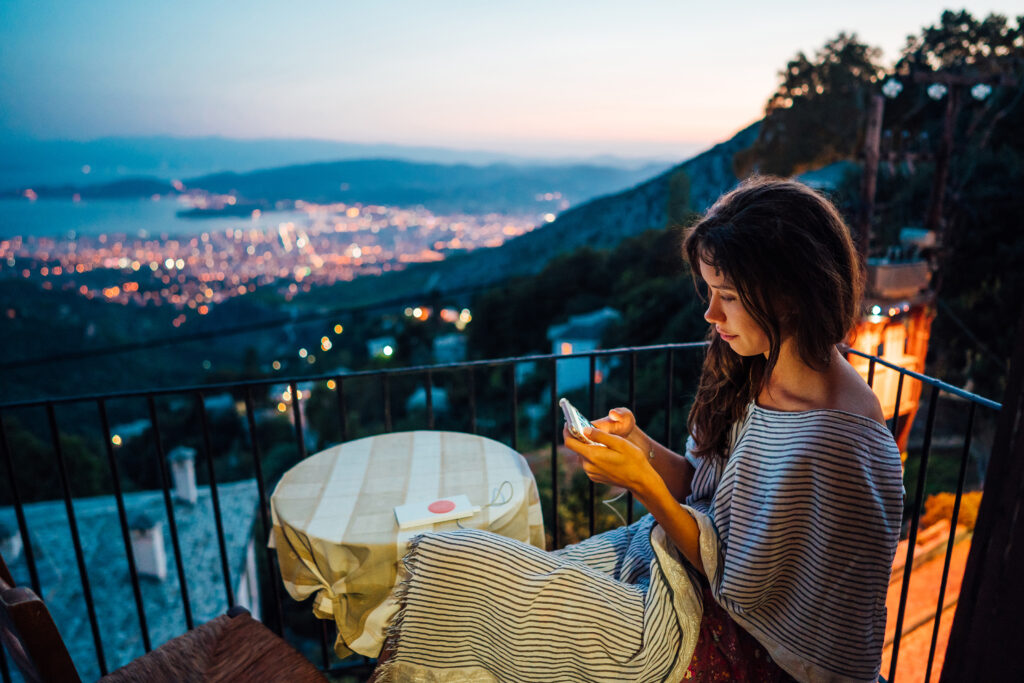 Solo Travel Italy: The girl sits on the balcony and looks into the smartphone, in the background the night city